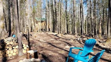 A forested area with a fire pit, stacked firewood, and blue Adirondack chairs.