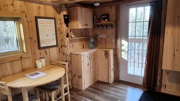 A wooden cabin interior with a dining table, chairs, a kitchen area, and a large window.