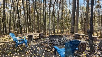 A forest setting with a fire pit, two blue Adirondack chairs, and a wooden bench.