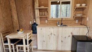 A wooden interior with a kitchen area including a table, chairs, a refrigerator, and a window with shelves above.