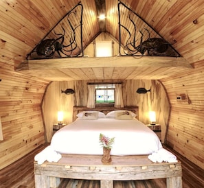 A cozy, wooden cabin bedroom with a bed, bedside lamps, a window, and a decorative fish on the ceiling.