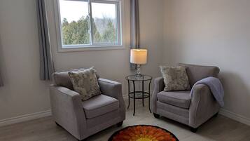 A room with two grey armchairs, a small round table with a lamp, a window with curtains, and a patterned rug.