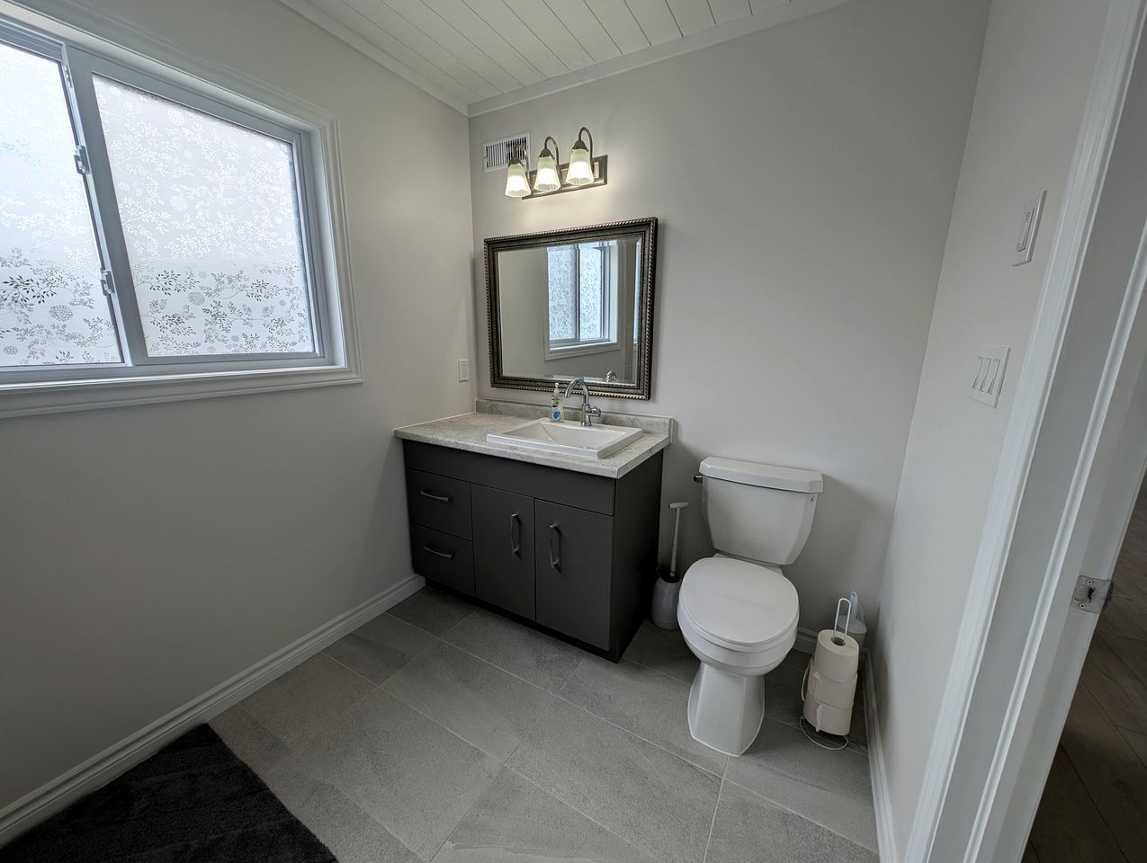 Une salle de bain avec des toilettes blanches, un meuble de couleur gris foncé, un lavabo blanc et une fenêtre en verre dépoli.