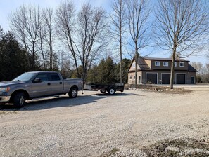 A gravel driveway with a pickup truck towing a trailer, a house with a garage, and leafless trees.