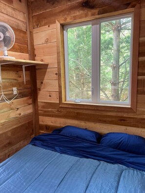 Une chambre dans un chalet en bois, avec un lit, une fenêtre donnant sur les arbres et un ventilateur fixé au mur.
