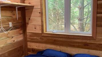 A wooden cabin room with a bed, a window with a view of trees, and a wall-mounted fan.