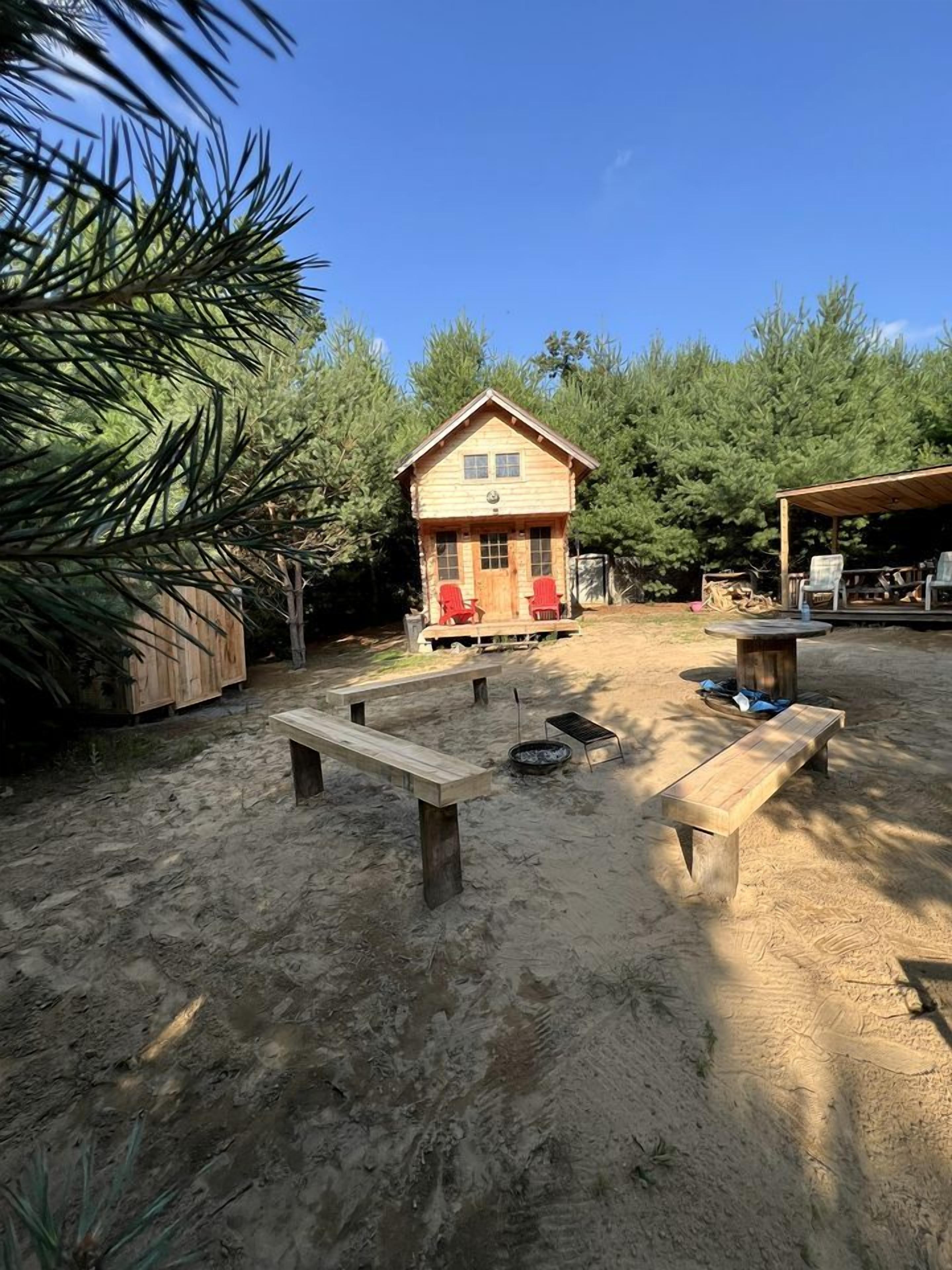 A wooden cabin with a porch, two red chairs, and a fire pit in a sandy outdoor area surrounded by trees.