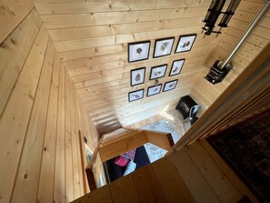 Ein Holzinnenraum mit schrägverdeckter Decke, gerahmten Bildern an der Wand, einem Kamin und einer Treppe.
