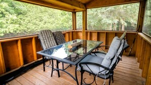 A wooden deck with a glass table and two chairs, surrounded by wooden railings and large windows. - Vintage Airtstream with Large Roofed Deck, BBQ, Fire Pit and Forest Views in Ontario (Blackstock)
