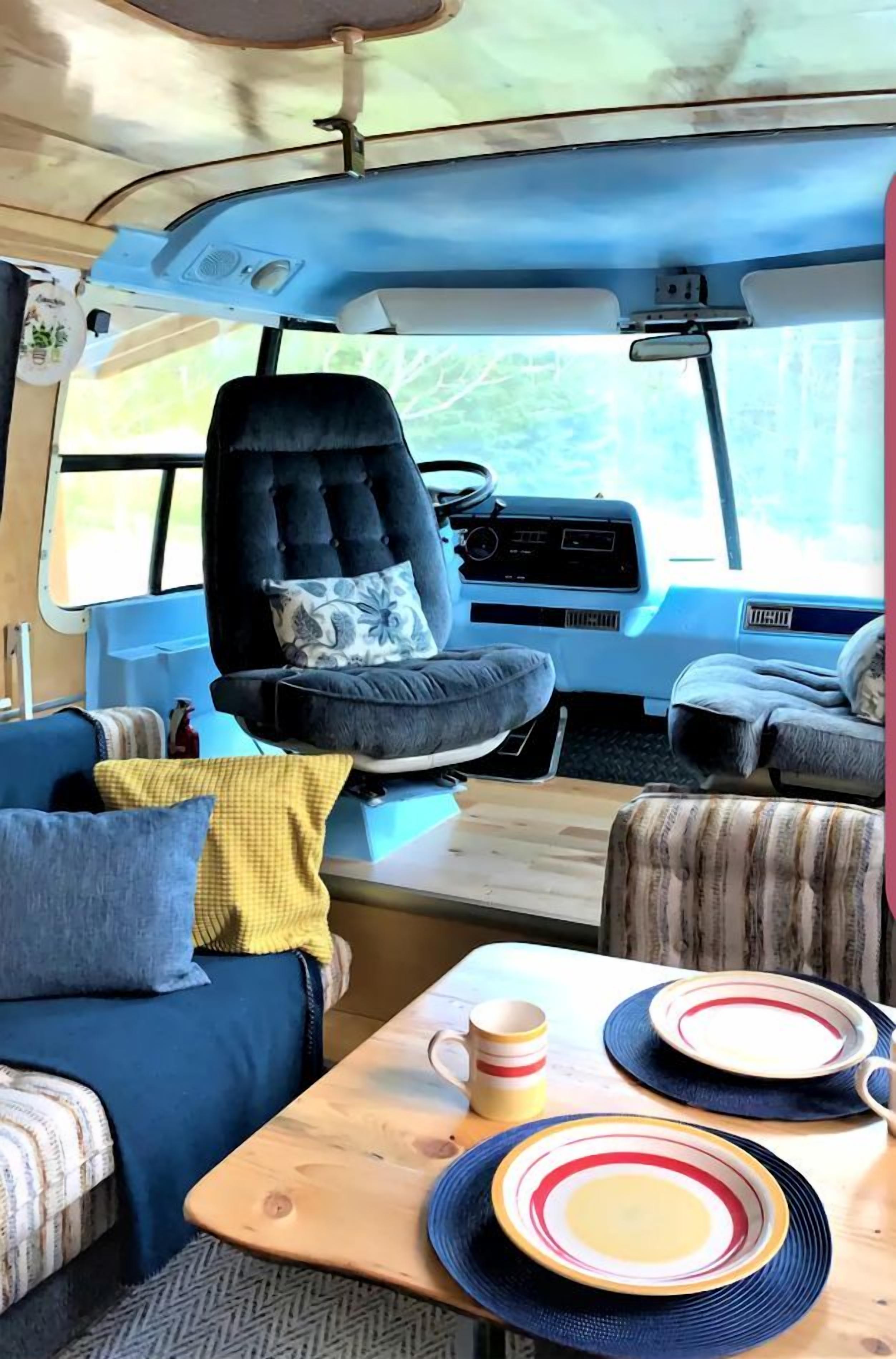 A cozy RV interior with a blue couch, a dining table with plates and a mug, and a black tufted chair.