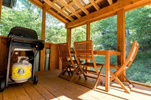 Een houten terras met een barbecue, propaantank, twee houten stoelen en een tafel.