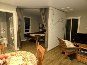 A room with a bed, a dining table with chairs, a small kitchen area, and a sliding glass door.