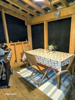 Een terras buiten met een tafel bedekt met een geruit tafelkleed, houten stoelen, een barbecue en een bloempot.