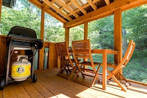 Une terrasse en bois avec un barbecue et une bouteille de propane, deux chaises en bois et une table.