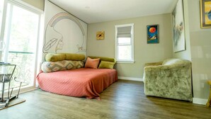 Un dormitorio con cama, una silla, un ventanal y obras de arte en las paredes.