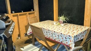 Une terrasse extérieure avec une table recouverte d’une nappe à motifs, des chaises en bois, un barbecue et une plante en pot.