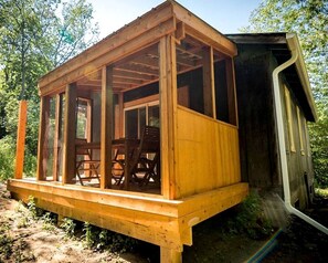 Een houten chalet met een overdekte veranda, een eethoek met stoelen, en een kleine aangebouwde schuur.