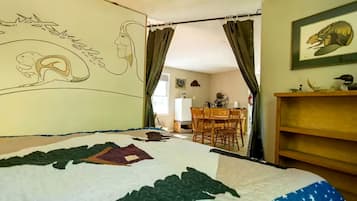 A bedroom with a bed, a wall mural, a dining area, and a kitchen in the background.
