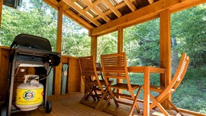 Une terrasse en bois avec un barbecue et une bouteille de propane, deux chaises en bois et une table.