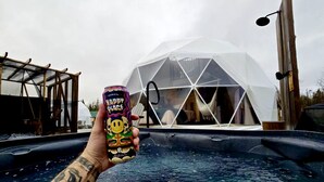 A hand holding a can with a colorful design, a transparent dome structure, and a hot tub in the foreground. - Cozy Dome Rental in Canada, for Romantic Weekends in Nature in Nova Scotia (Antigonish)