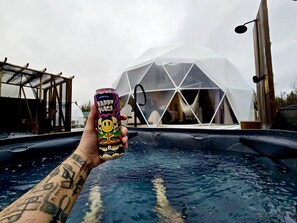 A hand holding a can with a colorful design, a transparent dome structure, and a hot tub in the foreground.