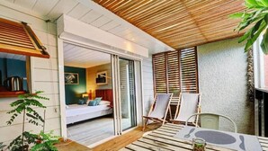 Een modern terras buiten met houten vloer, een eettafel en stoelen. Er is een schuifdeur naar een slaapkamer met een bed en nachtkastje.