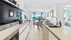 A modern kitchen with a built-in stove, a dining area with a table and chairs, and a view of the ocean through large windows. - Soul Surfers Paradise - Lvl11 Ocean View - Apartment (Surfers Paradise)
