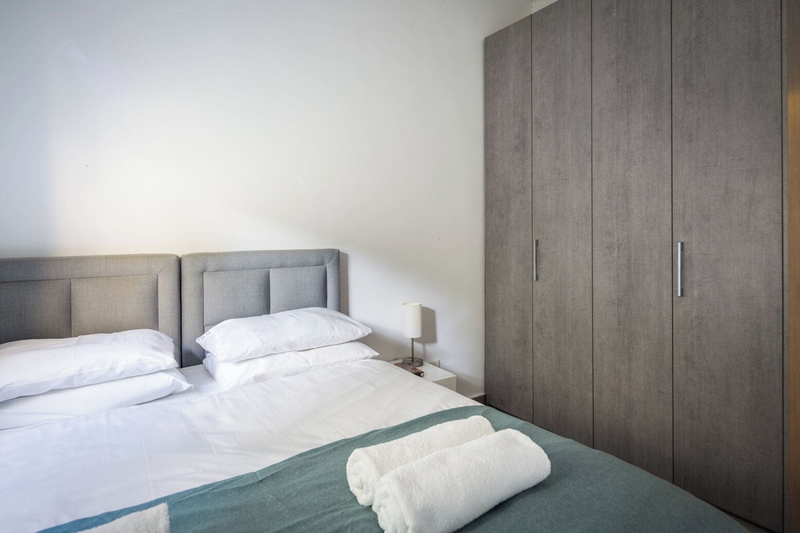 Un lit bien fait, avec du linge de lit blanc et un plaid vert, une tête de lit grise et une armoire en bois dotée de deux poignées.