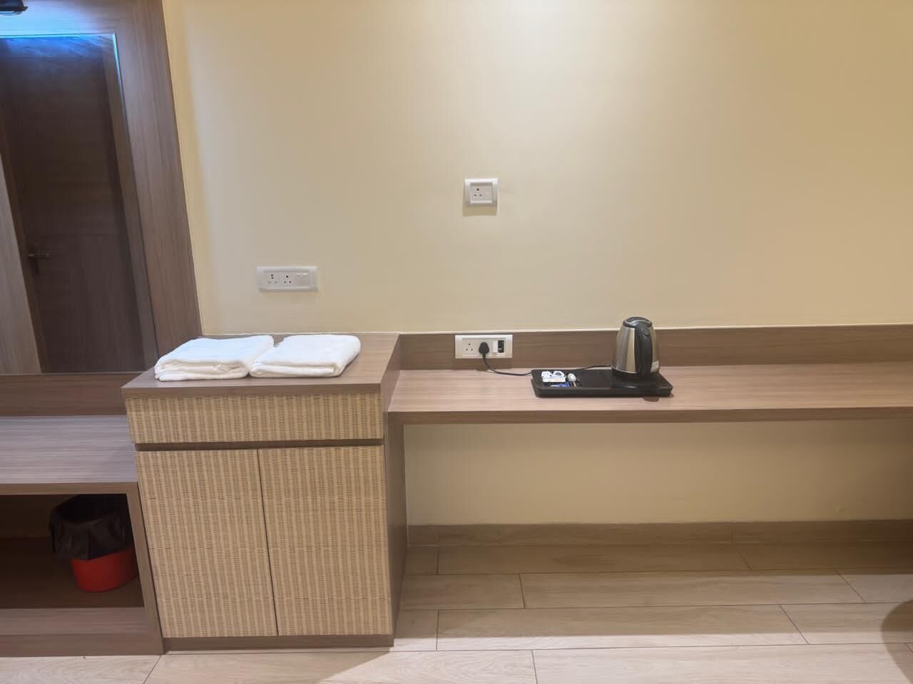 A hotel room with a wooden bench, a small wicker side table with towels, a kettle, and a red trash bin.