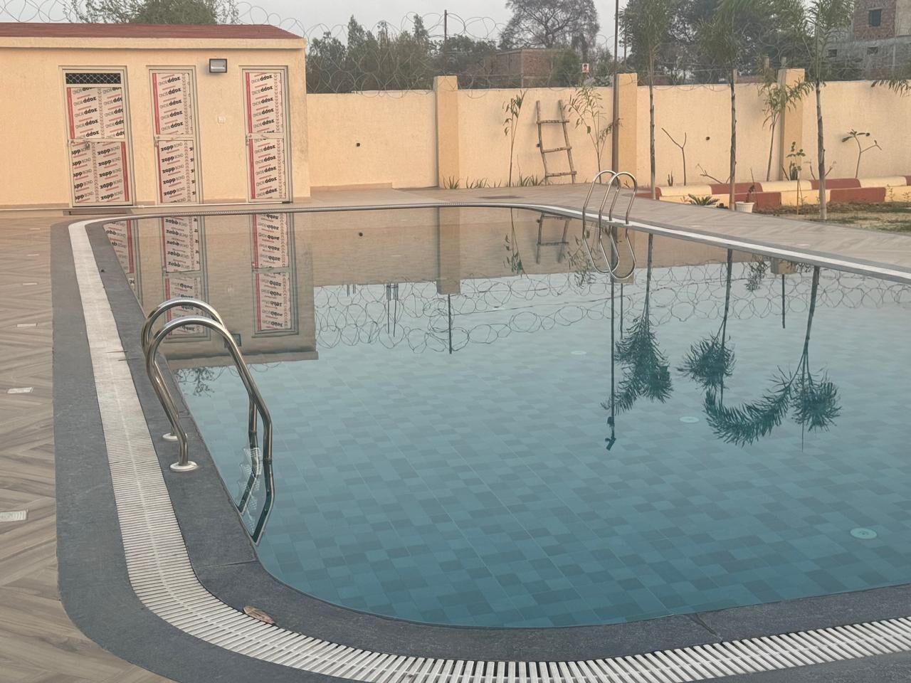 A swimming pool with a metal ladder, surrounded by a tiled deck and a low wall with a barbed wire fence.