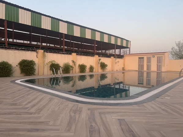 Une piscine dotée d’une terrasse carrelée, entourée d’un petit muret et bordée d’un bâtiment surmonté d’un auvent à rayures vertes et blanches.