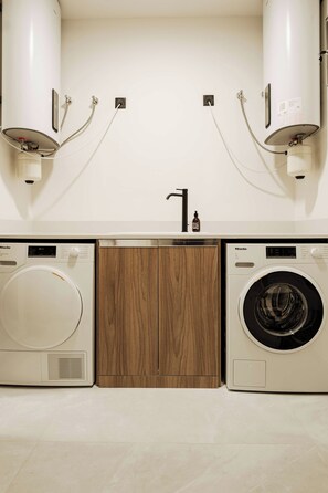 A modern laundry room with two front-loading washing machines, a wooden cabinet, and a sink with a faucet. - High-floor views, hot tub & sauna | AZ13 (Sliema)