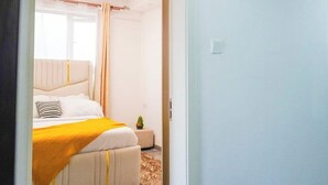 A bedroom with a bed, a small bedside table, and a window with curtains. - Cobot Suites Marcus Garden (Nairobi)