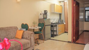 A compact living space with a sofa, a small round table, a heart-shaped chair, a compact kitchen with a refrigerator, microwave, and stove, and a wooden door leading to another room. - Cobot Suites Marcus Garden (Nairobi)