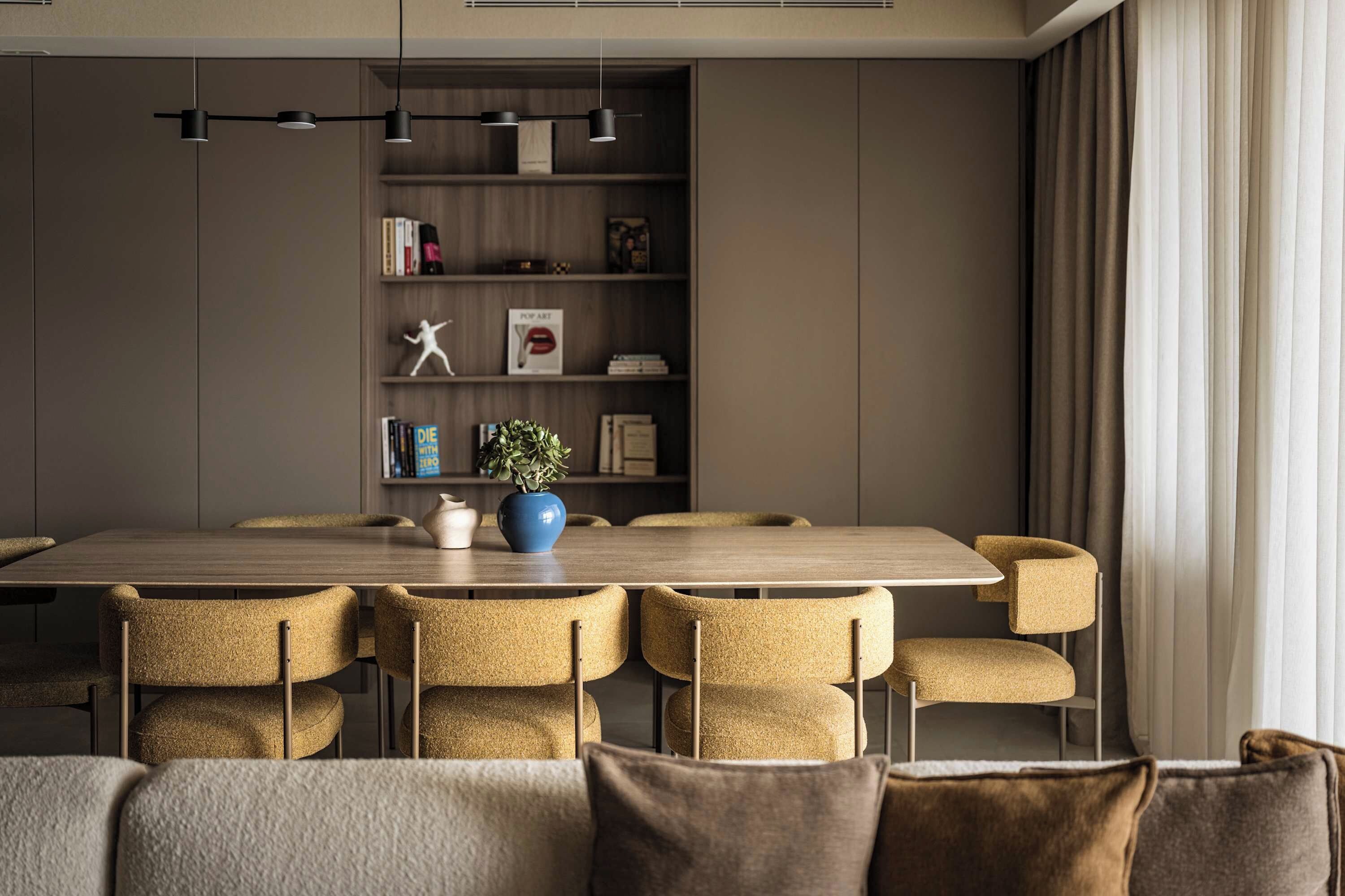 A modern dining area with a wooden table and six chairs, a bookshelf with books and decorative items, and a potted plant on the table.
