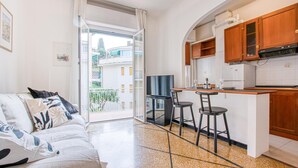 A compact living space with a kitchenette, a sofa with patterned cushions, a dining area with two stools, and a sliding glass door leading to a balcony. - Lovely apartment in Rapallo with WiFi (Rapallo)