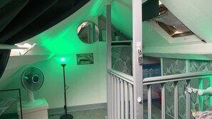 Una habitación con techo inclinado, un ventilador de pie, una lámpara con luz verde, un certificado enmarcado y una cama con almohada estampada con flores.
