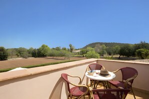 Un balcón con sillas de mimbre, una mesa y vistas a un jardín ajardinado con árboles y una colina.