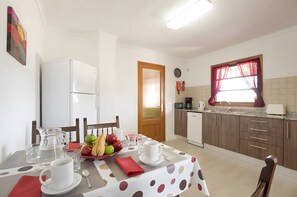 Un comedor con una mesa dispuesta para dos personas, un refrigerador, una cafetera y una ventana con cortinas.