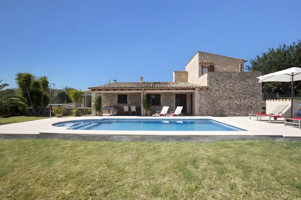 Una casa de dos pisos con piscina, mobiliario exterior y un muro de piedra.