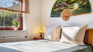 Ein Schlafzimmer mit einem Bett, Kissen, einem Nachttisch mit Lampe, einem Fenster mit Jalousien und einem Wandbild.