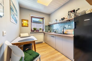 A compact kitchen with a refrigerator, sink, and wooden countertop. There is a window with a view of the outside, a chair, and a table with a cushion.
