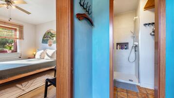 Een kamer met een bed, een plafondventilator, een kapstok aan de muur, een douche en een raam met jaloezieën.