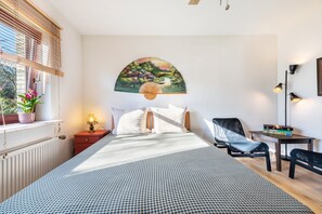 Ein Schlafzimmer mit Bett, Nachttisch, Lampe, Stuhl, Schreibtisch und Wandventilator.