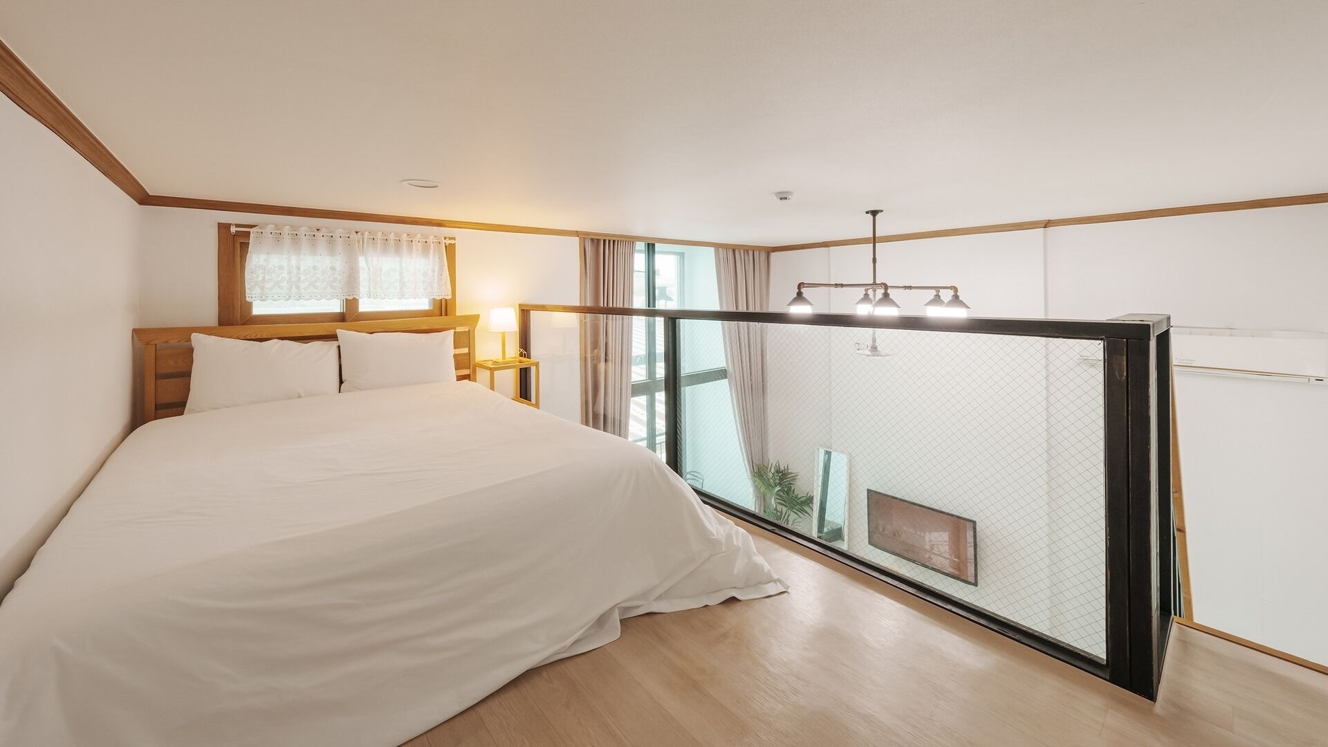 Un dormitorio con cama, mesita de noche, ventana con cortinas, luz de techo y vista a través de un panel de vidrio.