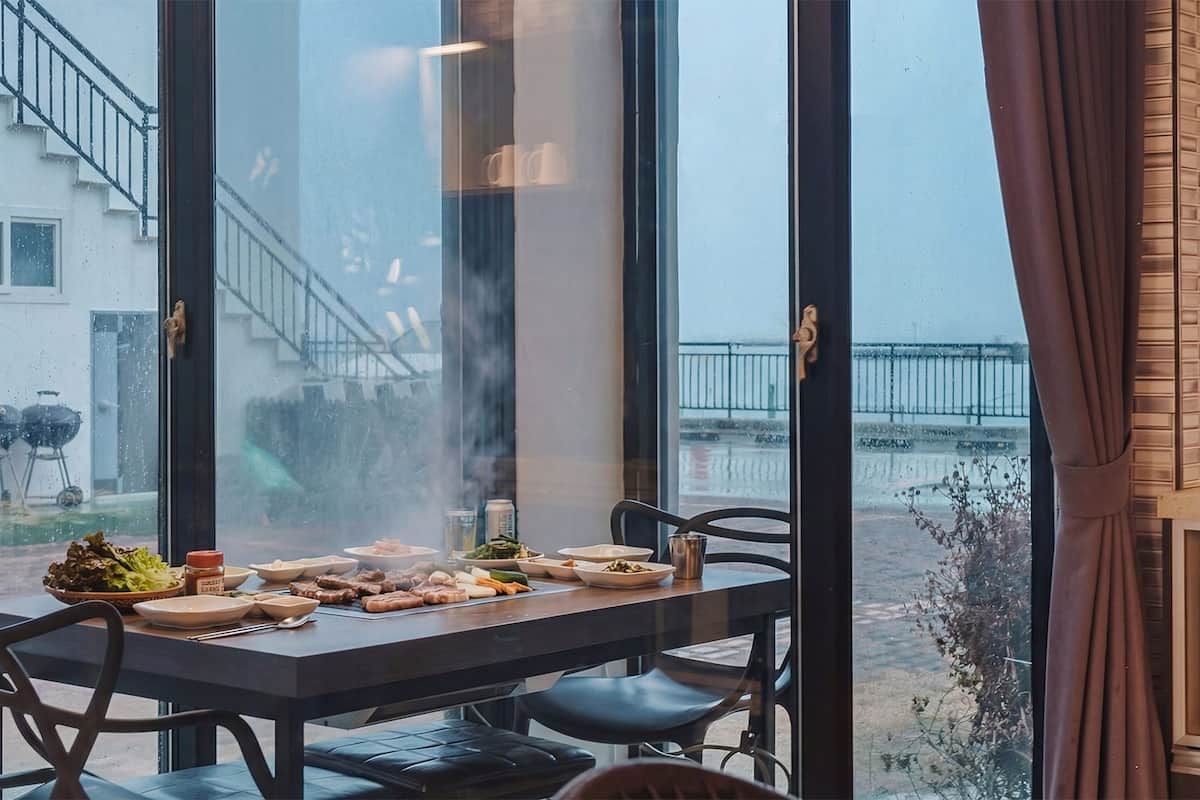 Un espace repas avec une table dressée pour le repas, de grandes fenêtres et une vue sur un bâtiment doté d’un escalier.