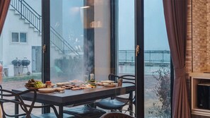 Un espace repas avec une table dressée pour le repas, de grandes fenêtres et une vue sur un bâtiment doté d’un escalier.