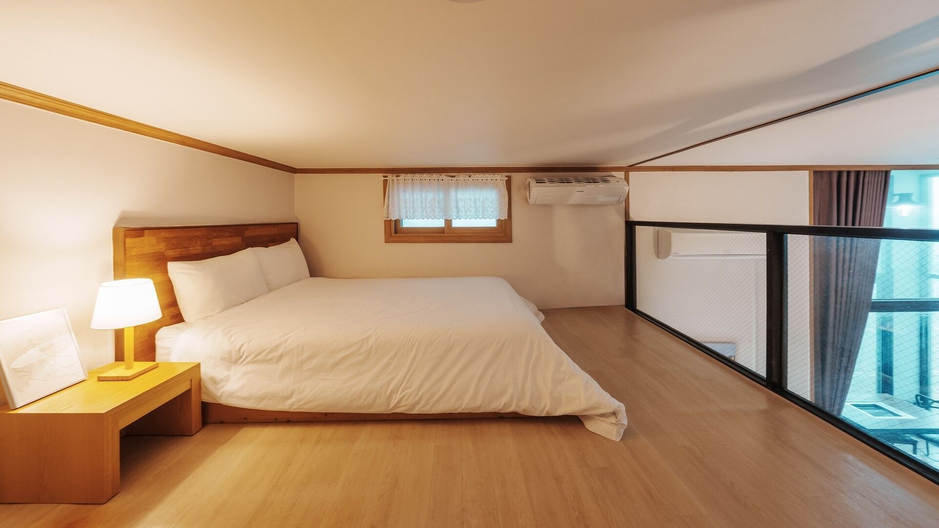 A hotel room with a bed, bedside table, lamp, window, air conditioning unit, and a balcony with a railing.