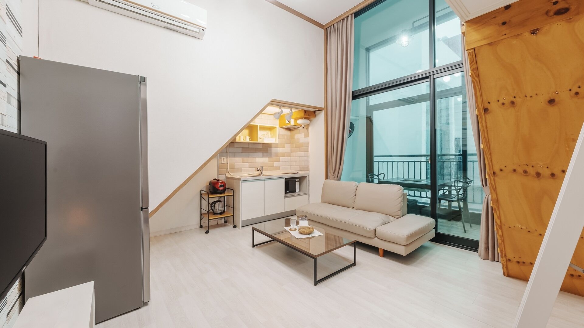 A modern apartment with a kitchenette, a sofa, a coffee table, and a large window with a balcony.