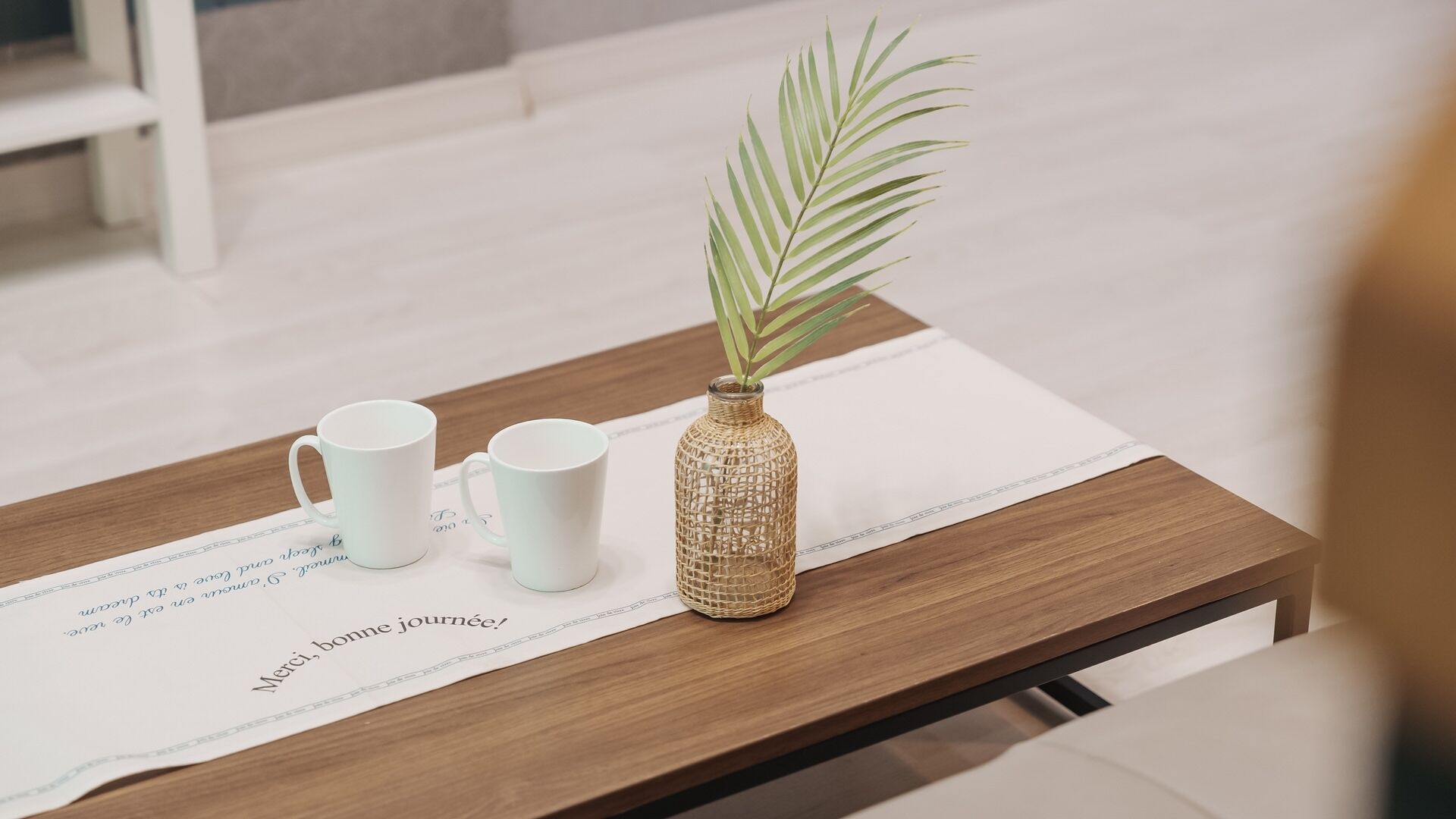 Una mesa de madera con un mantel individual blanco, dos tazas de cerámica blanca y un florero de vidrio texturizado con una planta verde.